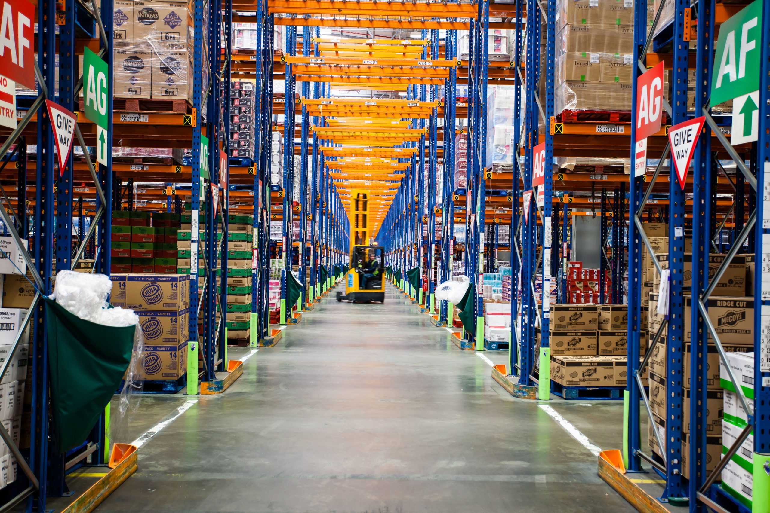 A lift truck travels up a long warehouse aisle with stocked racking either side